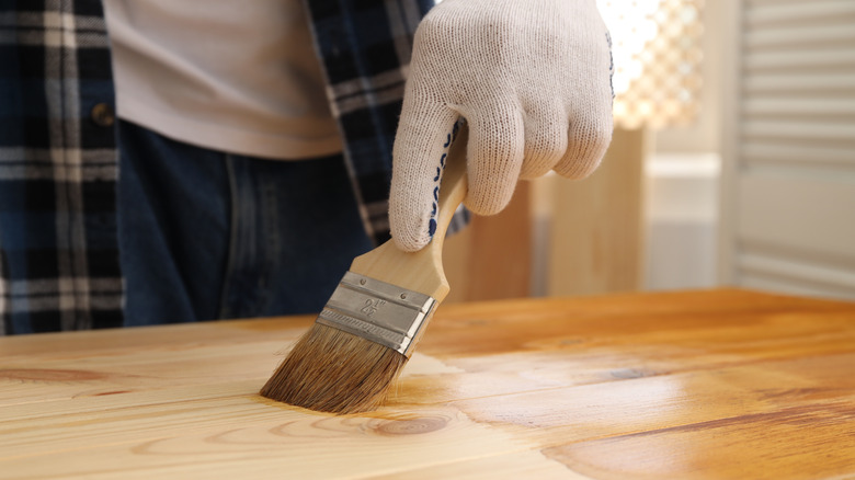 A gloved hand applies stain to wood with a brush.