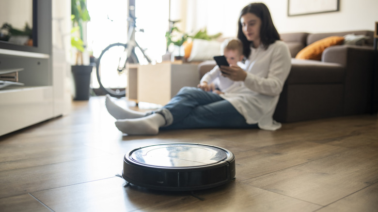 A woman and child sit on a wood floor while a robot vacuum operates