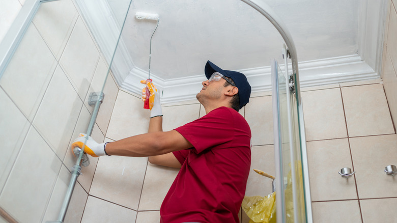 A man paints a bathroom while wearing a hat and protective eyewear.
