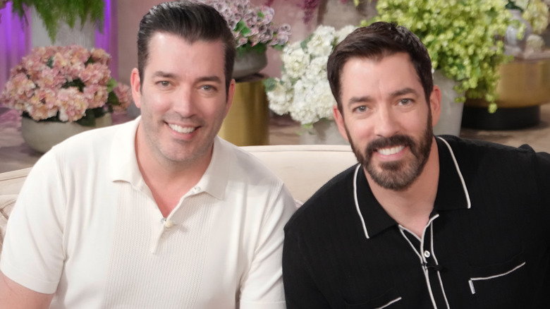 Jonathan and Drew Scott sitting side by side on a couch and smiling at the camera.