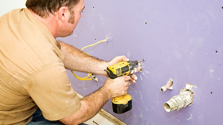 A person drilling into purple drywall with two pipes sticking out of the wall next to them