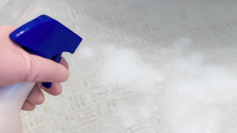 Gloved hand spraying a cleaning solution on white-tiled bathroom floor