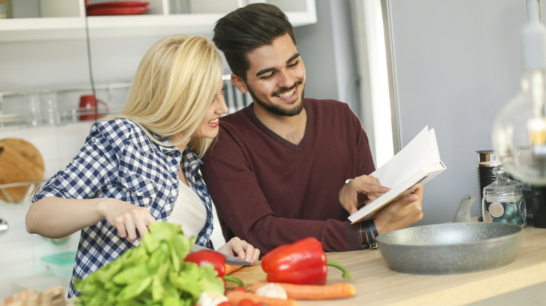 Couple peers over cookbook as they cook a meal in the kitchen