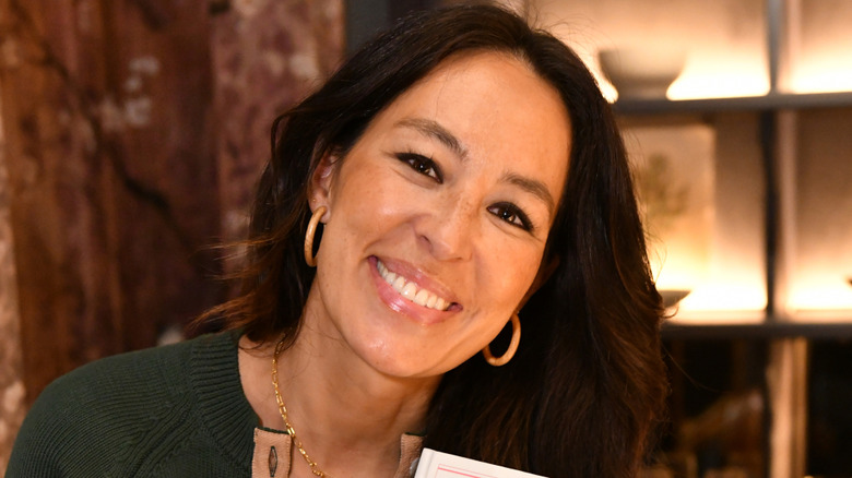 Joanna Gaines smiling with long dark hair wearing gold hoop earrings, photographed indoors with warm lighting and shelves in the background.