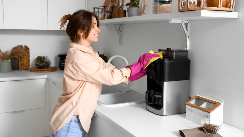 A woman wears gloves to clean a coffee maker in a bright, white kitchen.