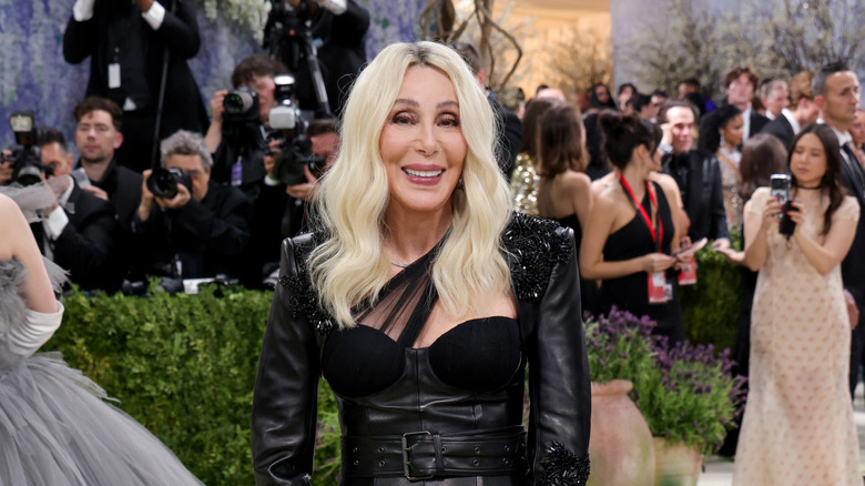 Cher in blond hair and a black leather outfit, smiling on the carpet at the Met Gala
