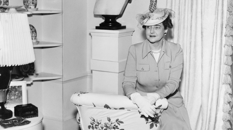 Dorothy Draper sits on the edge of an armchair in a skirt suit and hat.