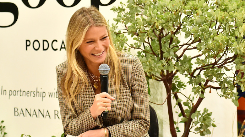 Gwyneth Paltrow in a plaid suit jacket, laughing into a mic at a Goop event