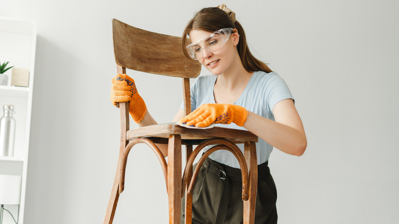 A woman in safety glasses sanding a vintage wood chair