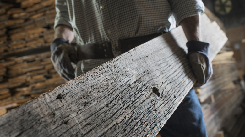 A man in gloves carries a large, old 2x4 of lumber into a construction site.