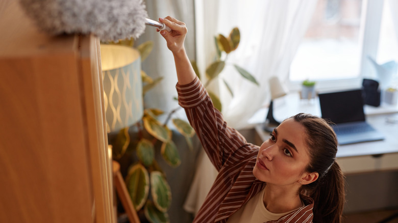 Young woman dusting tall shelves with duster