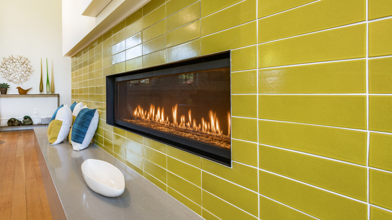 A tiled, chartreuse backsplash surrounding a rectangular, lit fireplace and accented with several pillows.