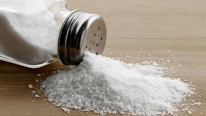 Salt spills out of a glass and metal salt shaker onto a wooden tabletop.