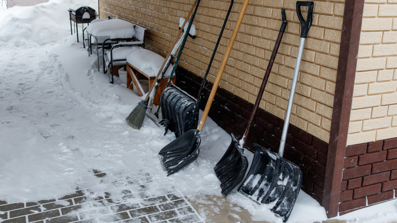 Multiple snow shovels of different sizes and weights leaning against a house with snow on the ground