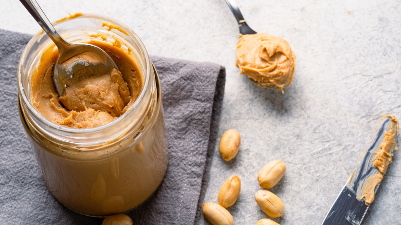 A spoonful of peanut butter in a jar of peanut butter, with a glass bowl of peanuts nearby