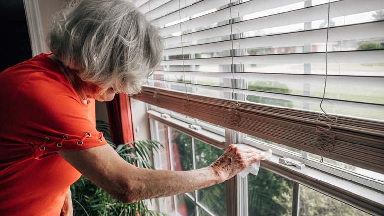 An older adult cleans the window frame of a window dressed with Venetian blinds.