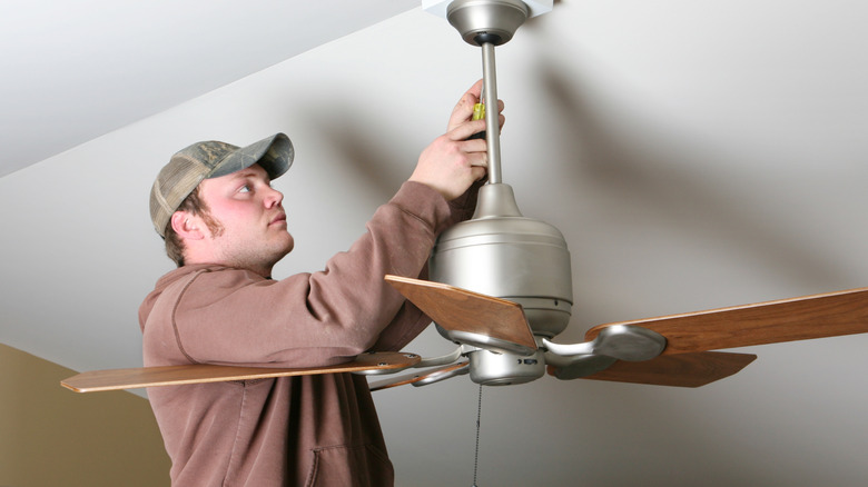 A man wearing a camo hat unscrewing an outdated ceiling fan from vaulted ceiling.
