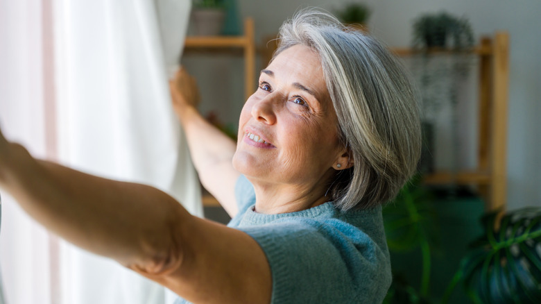 Woman opening white curtains with wood shelves in the background