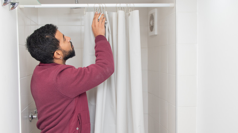 A man hangs up a plain white shower curtain in an all-white bathroom.