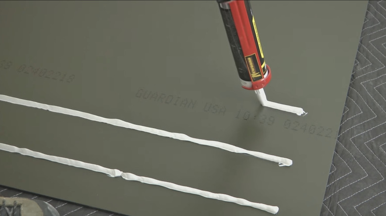 A person uses a cartridge gun to apply thin strips of adhesive to the back of a bathroom mirror