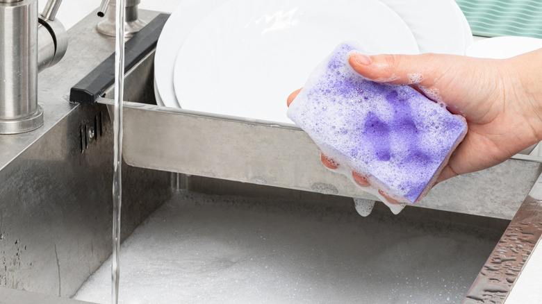 A person holds a sudsy sponge over a sink of soapy water and dishes