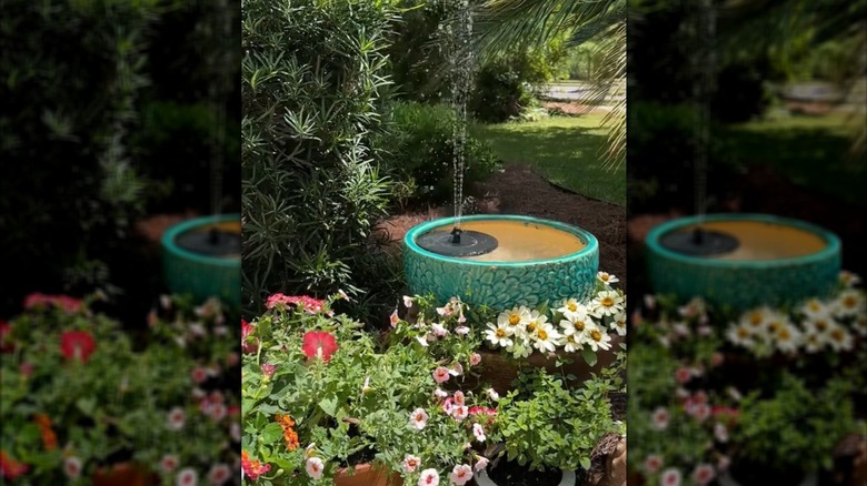 A green pot with outside floral designs is buried in a planter, filled with water and a solar-based fountain accessory.