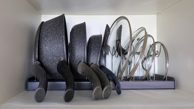 A bunch of pots, pans, and lids stacked neatly in a storage rack inside a kitchen cupboard.