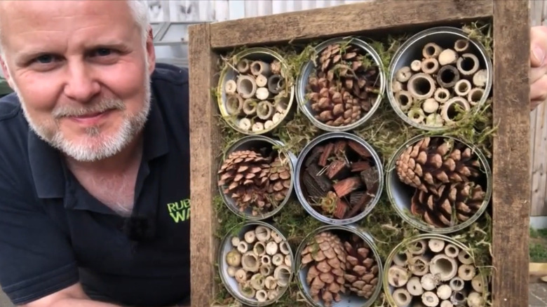 Man displaying a bug hotel with several tin cans inside a frame with various materials for bugs