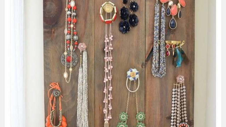 A wood-backed, wall-hung jewelry organizer that features cabinet pulls with necklaces hanging off it.