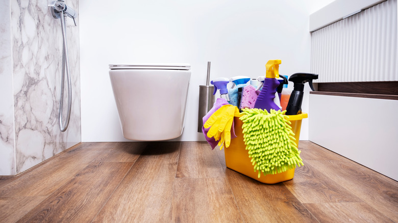 A yellow bucket filled with cleaning solution sprays, gloves, rags, sponges, and more next to a toilet