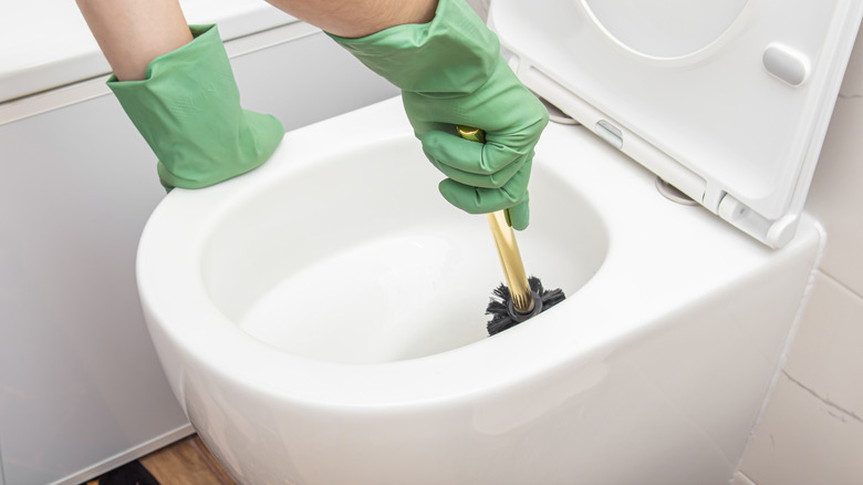 A person wearing green rubber gloves cleans a toilet bowl using a toilet brush