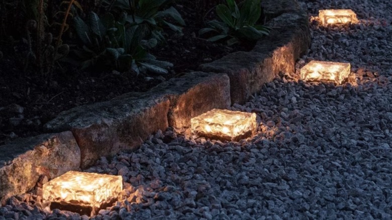 Four glass cubes emitting a soft, warm glow sit on top of gray gravel, next to a curved stone border.