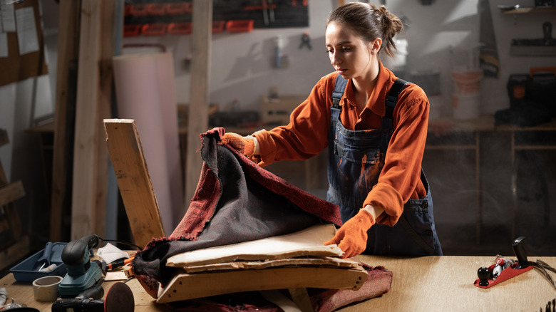 Female craftsman in overalls reupholstering vintage wooden chair.
