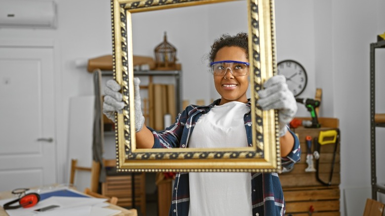 Woman in work glasses and gloves holding thrifted picture frame in garage