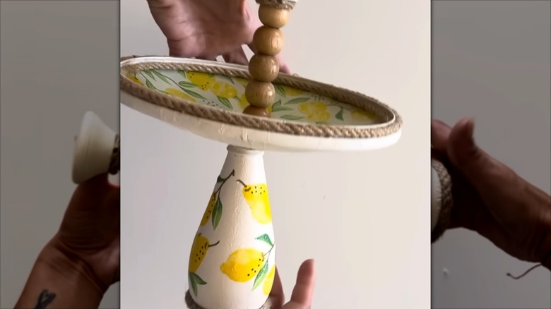 A pair of hands holds a decorative stand made from a pot lid and a glass bottle.