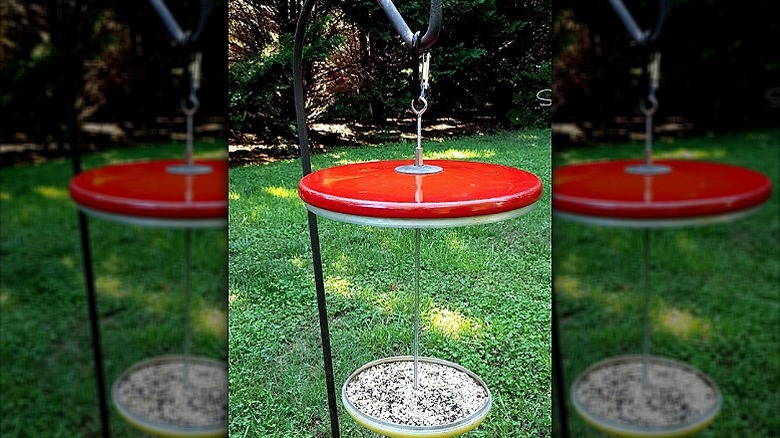 A bird feeder made using two pot lids hanging on a shepherd's hook in a garden.