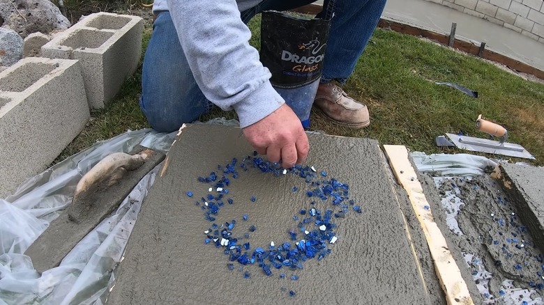 A person kneeling down next to a concrete paver and sprinkling shards of blue glass in a circle shape