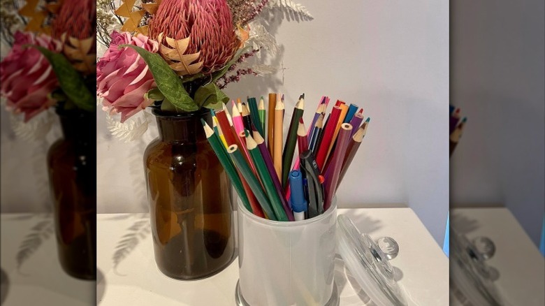 A candle jar repurposed into color pencil holder next to vase of flowers
