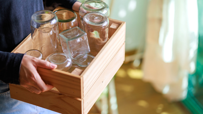 A person's hands are holding a wooden crate full of empty glass jars.