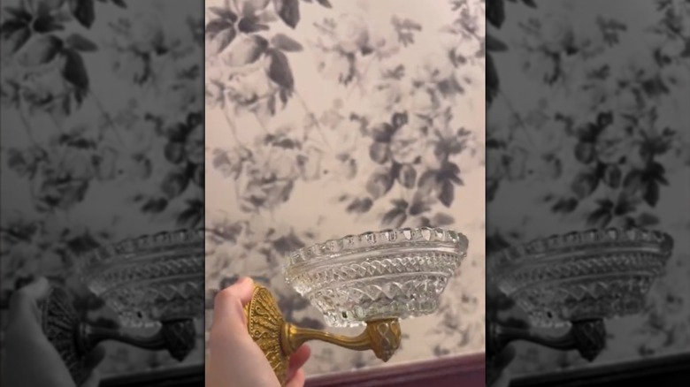 A hand holds a DIY soap holder made from a glass candle holder and a brass hook.