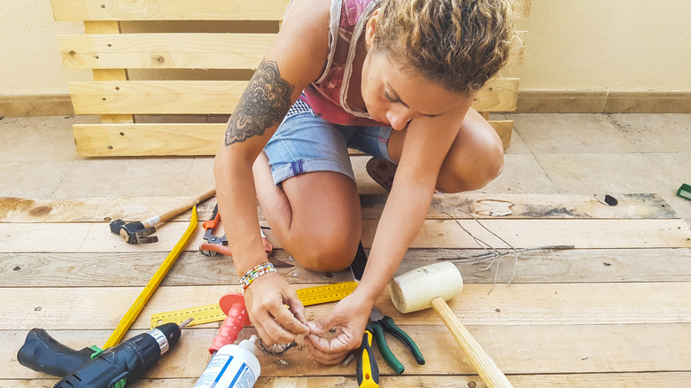 A women sits on a deck surrounded by carpentry tools with a wooden pallet behind her.