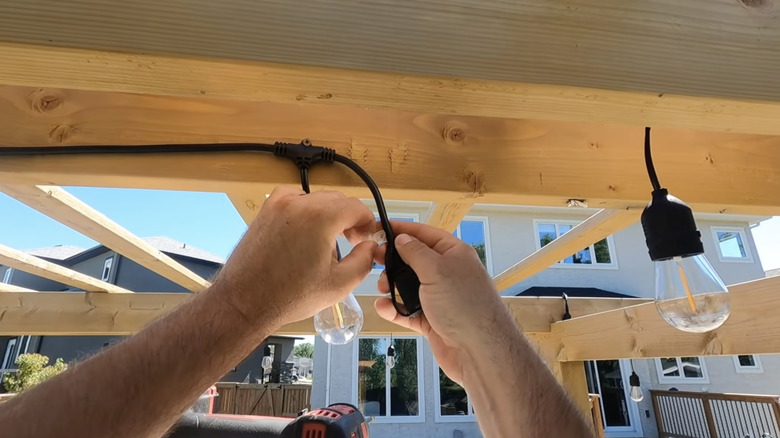 A person hangs string lights with large Edison bulbs to a wooden pergola roof.