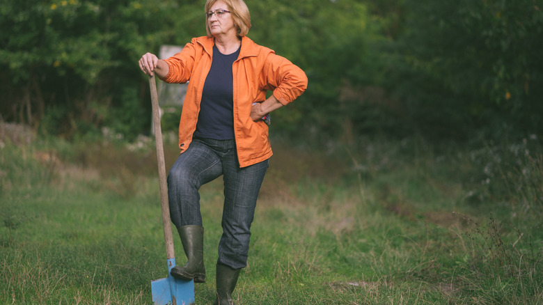 A woman in an orange jacket stands in her backyard with one foot on a shovel.