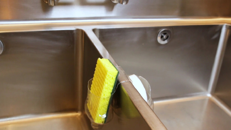 A sponge holder made from a bottle draped over a center divider in a steel sink.