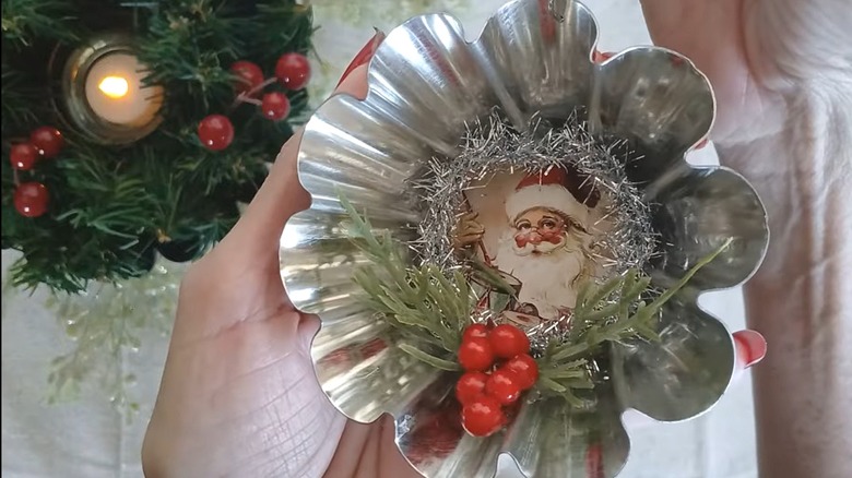 Person holding a finished tart pan ornament with a vintage Santa image, tinsel, greenery, and berries