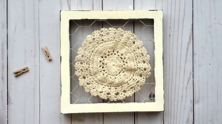 A woman demonstrating how she made a doily wall decor piece from a Dollar Tree chicken wire picture frame with tiny clothespins.