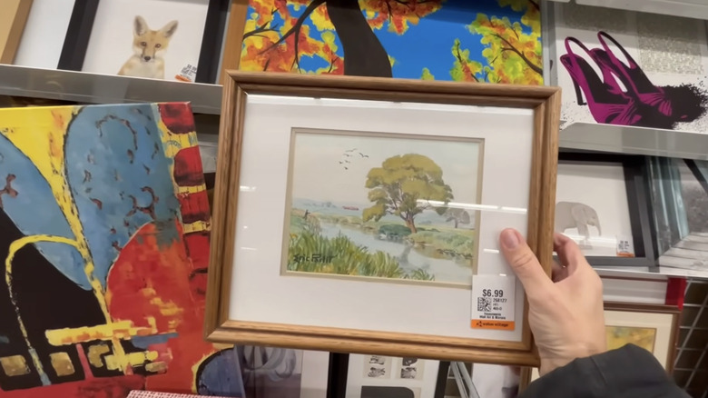 A hand holding a small painting with other paintings behind it.