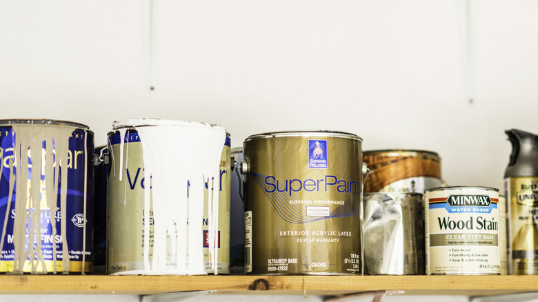 A horizontal shot of a collection of opened and unopened American brand cans of paint, wood stain and paint sprays organized neatly on a wooden shelf.