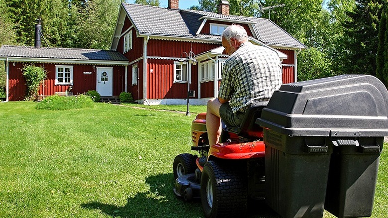 A person looks down at grass while riding a lawn mower in front of a red and white house