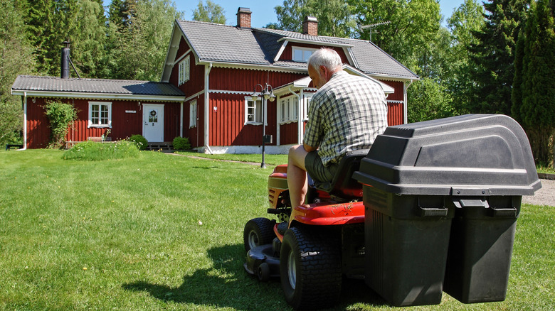 Older man on a red riding lawn mower going toward a red and white house.